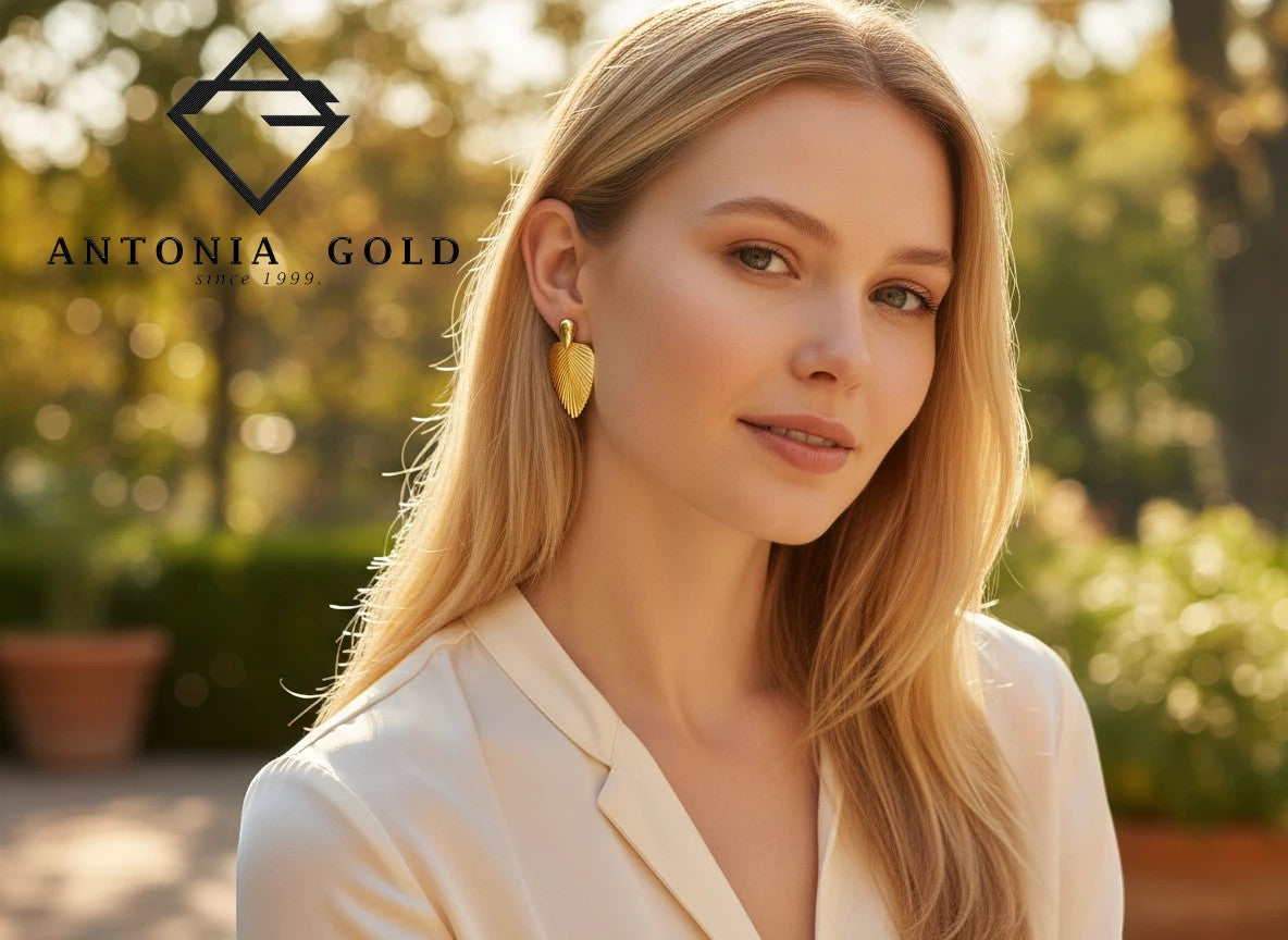 Cercei Antonia Gold în formă de frunză aurie cu striații fine, purtați de o femeie blondă într-un cadru natural luminos.